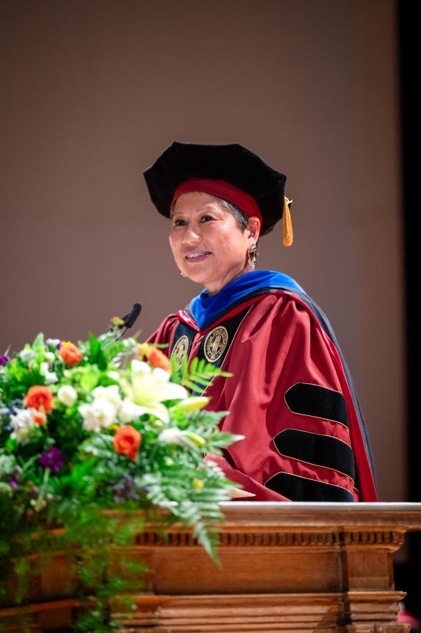 Dean Marianne Yoshioka wearing red academic robes and cap stands at a podium full of flowers and greenery