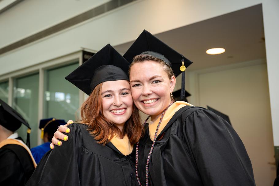 Two students wearing black academic robes, caps and yellow master's hoods smile and hug