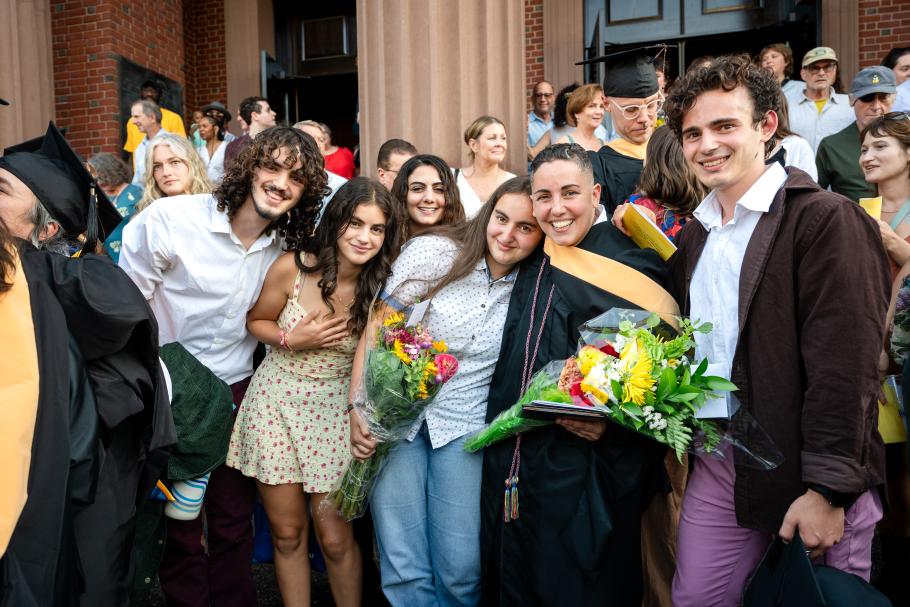 Lev Fein wearing black academic robes and yellow master's hood holds a bouquet hugging their four children who are standing around them. Behind are other grads and families and the red pillars of John M Greene Hall