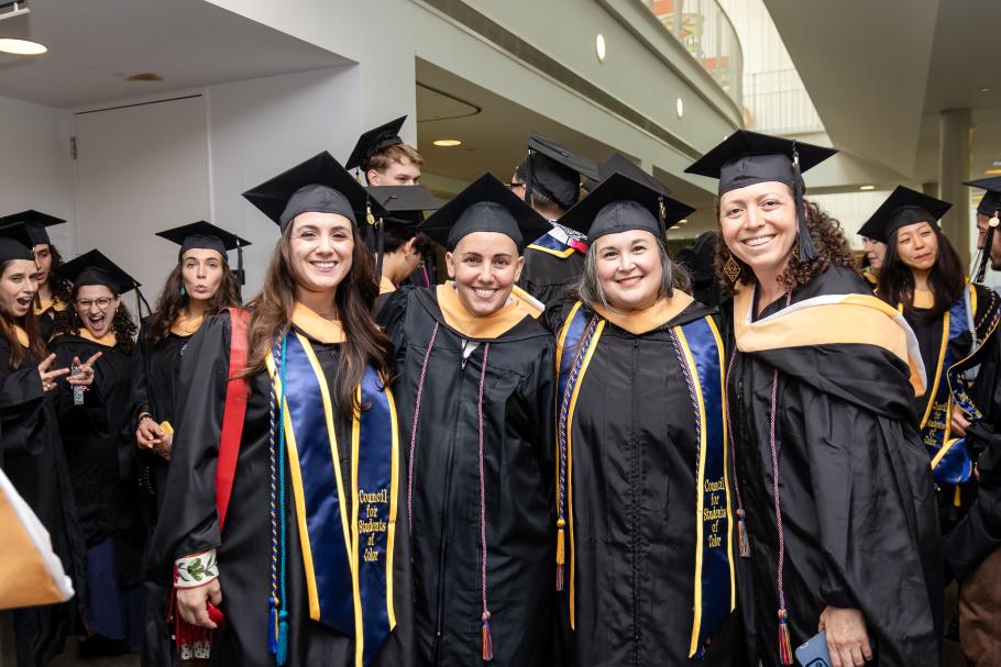 Four graduates wearing black academic robes and caps stand in a row hugging and smiling. Behind are other grads and white walls of the Campus Center.