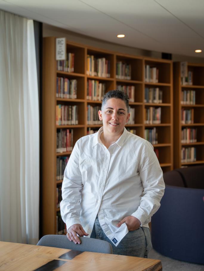 Lev Fein wearing white button down stands behind a wooden table with bookshelf behind in Neilson Library