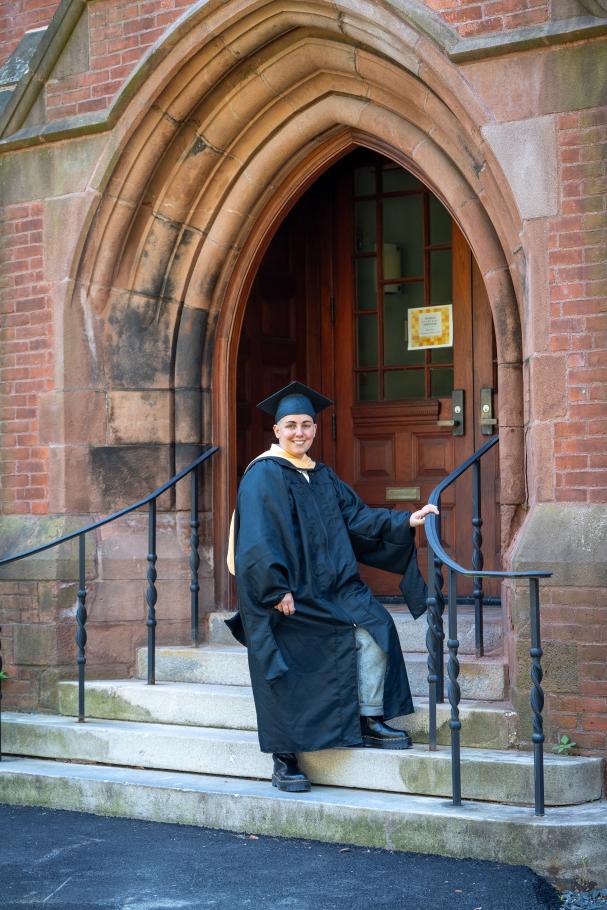 Lev Fein wearing black academic robes and cap stands on cement steps with brick arch above