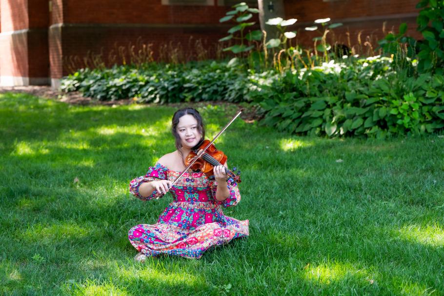 Maryann Han sits in the grass playing her violin.