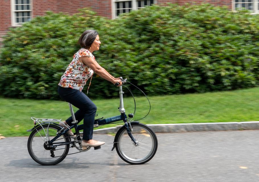 Marianne Yoshioka rides a bike on an asphalt path with greenery and brick buildings behind in 2025