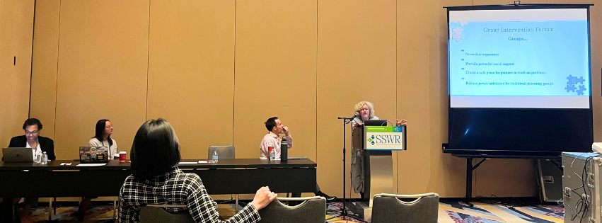 Marsha Pruett stands behind podium that reads SSWR with three people sitting to her left and a large presentation screen