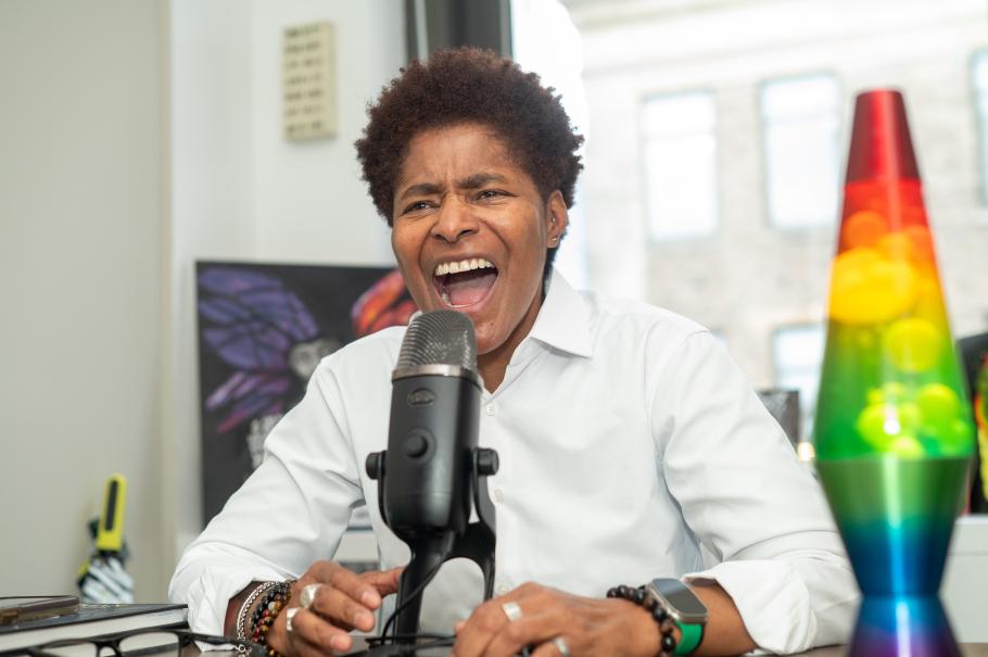 JD Fuller sits behind a mic with a rainbow lava lamp and window behind, laughing
