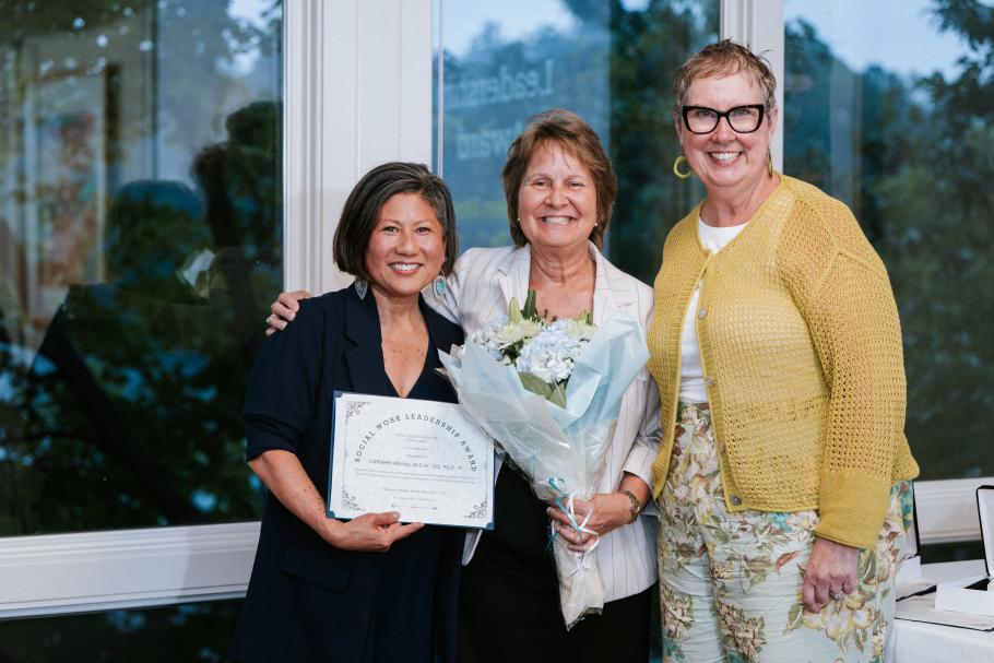Dean Marianne Yoshioka holds an award and stands with Cathleen Morey, award winner who is holding a bouquet of flowers and Dawn Faucher at the 2025 alumni awards. Glass windows in background.