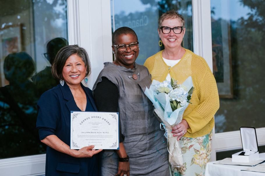 Dean Marianne Yoshioka holds award next to Lisa Moore and Dawn Faucher who holds bouquet at 2025 Alumni Awards. Large floor-to-ceiling windows behind.
