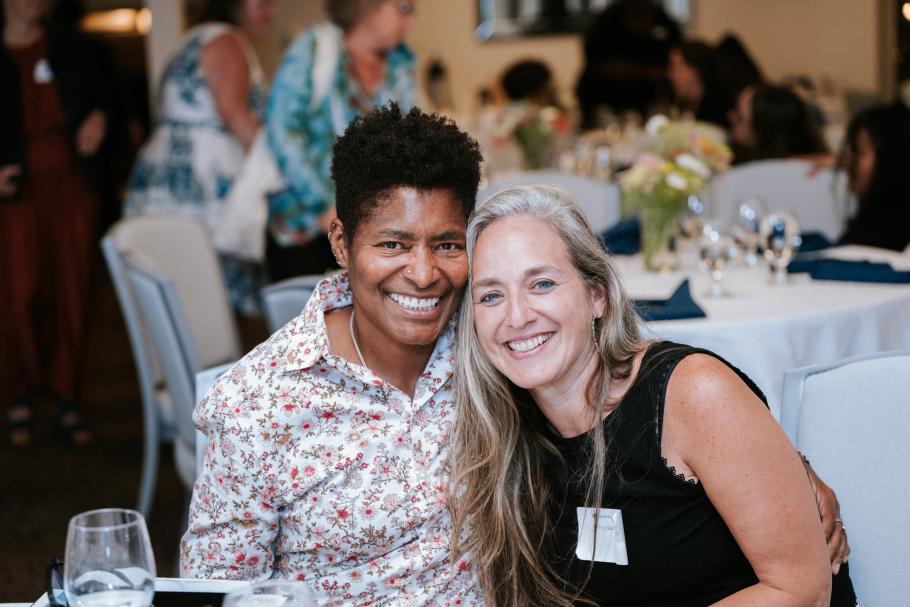 JD Fuller and her partner sit with arms around each other at a table during the 2025 Alumni Awards. Behind are tables with white tablecloths, people, flowers