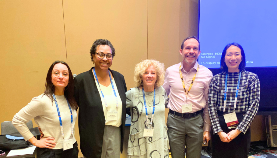 Five people including Brandyn-Dior McKinley and Marsha Pruett stand in a row with arms around each other with tan wall and large projector screen behind