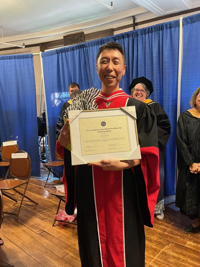 Kenta Asakura wearing academic red and black robes holds 3rd annual Laura Rauscher Memorial Award 