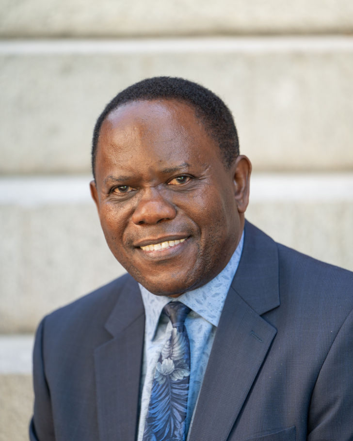 Hugo Kamya smiling in blue suit and tie with grey cement wall behind