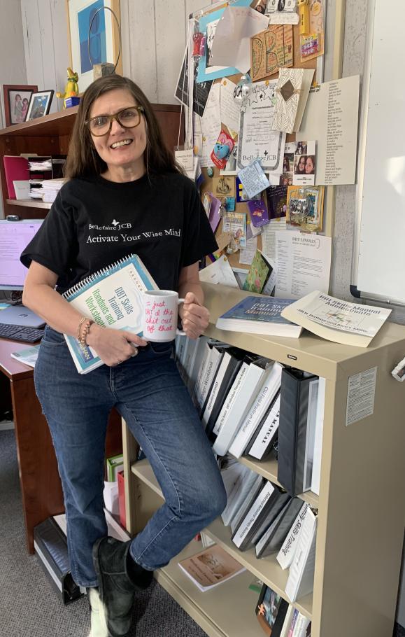 Samantha Mishne holding mug and folder, leaning against bookshelf