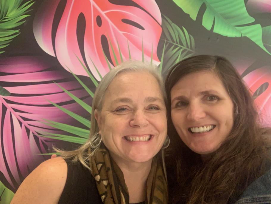 Denise Balzer and Samantha Mishne smiling in front of black, pink and green leaved wallpaper