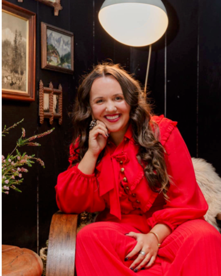M Barney wearing red dress sitting in chair with art on dark wall behind and round lamp above