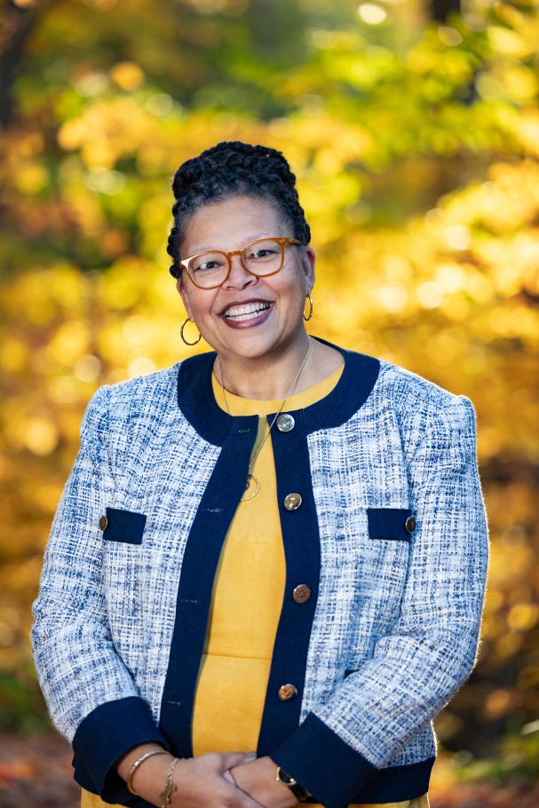 Sarah Willie-LeBreton smiles toward the camera in front of fall foliage, wearing a yellow shirt with a blue jacket on top