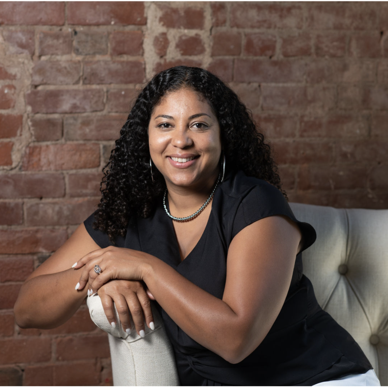 Photo of a person with long curly dark hair sitting on a white tufted couch. They are wearing a black v-neck t-shirt and have their arms resting on the arm of the sofa.