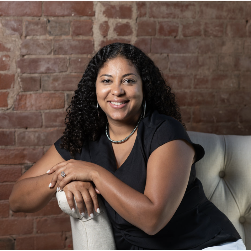 Photo of a person with long curly dark hair sitting on a white tufted couch. They are wearing a black v-neck t-shirt and have their arms resting on the arm of the sofa. 