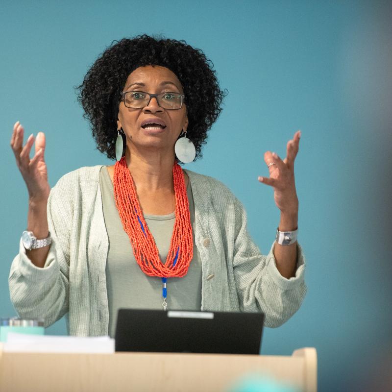 A presenter stands at a podium. They are using their hands as they speak. They are wearing an orange statement necklace and tan sweater and have shoulder length hair.