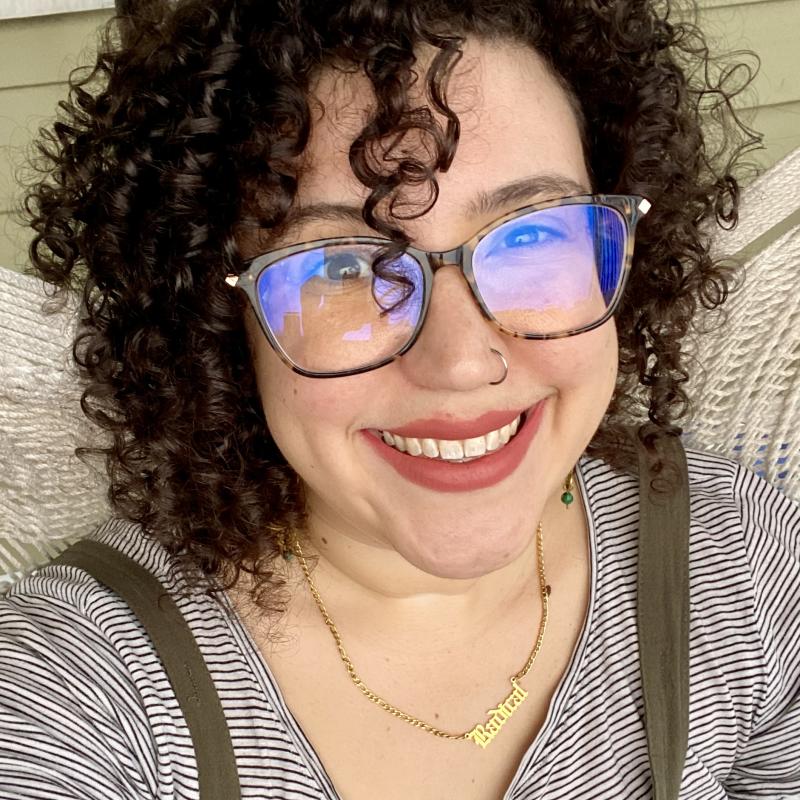 Selfie of Hannah Biskind, wearing glasses and a striped shirt sitting on a white wicker seat