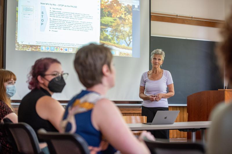 Three students sit in a classroom facing away from the camera. A professor stands at the front of the room in front of a large screen with course content projected on it.