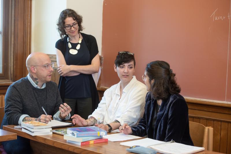 A professor stands next to three students who are seated at a table. The professor is listening to the students who are engaged in conversation. There are books and papers all over the table. 