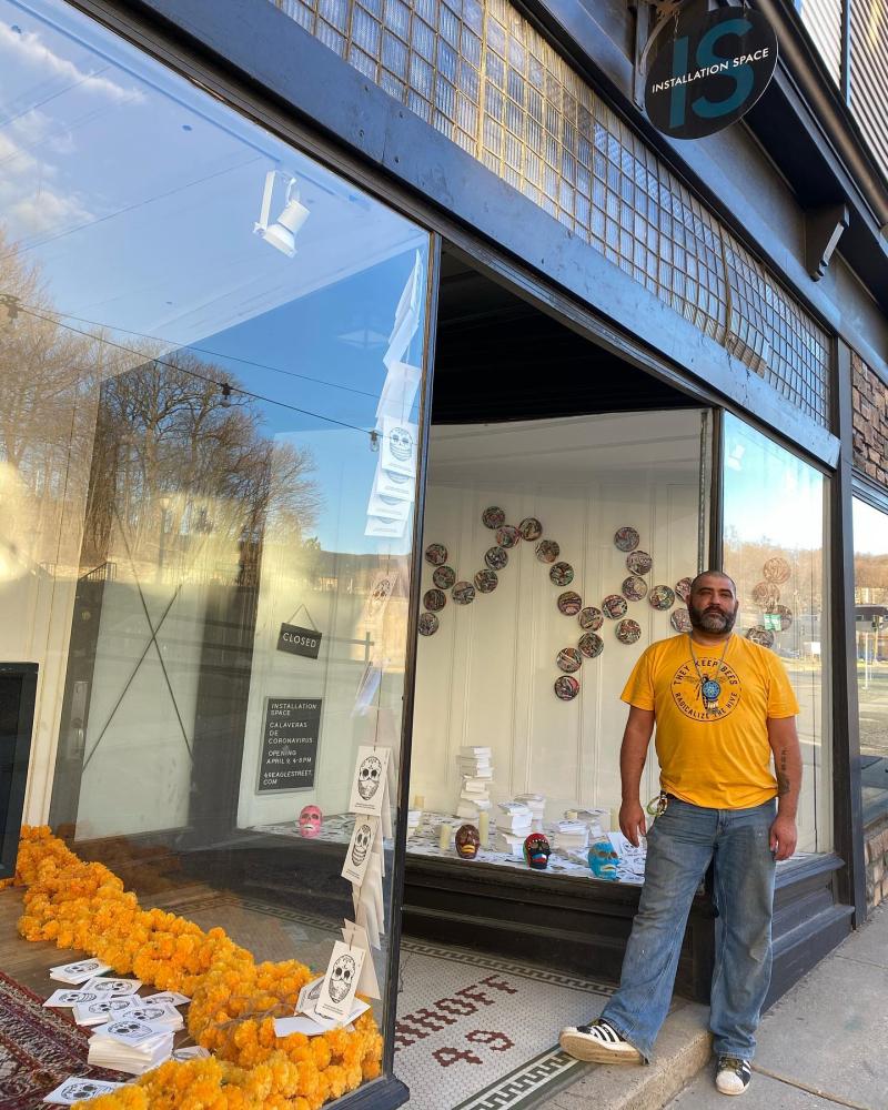 A person with a beard and short hair wearing a yellow shirt, jeans, white sneakers stands outside glass storefront with marigolds/cempasuchil in the windows
