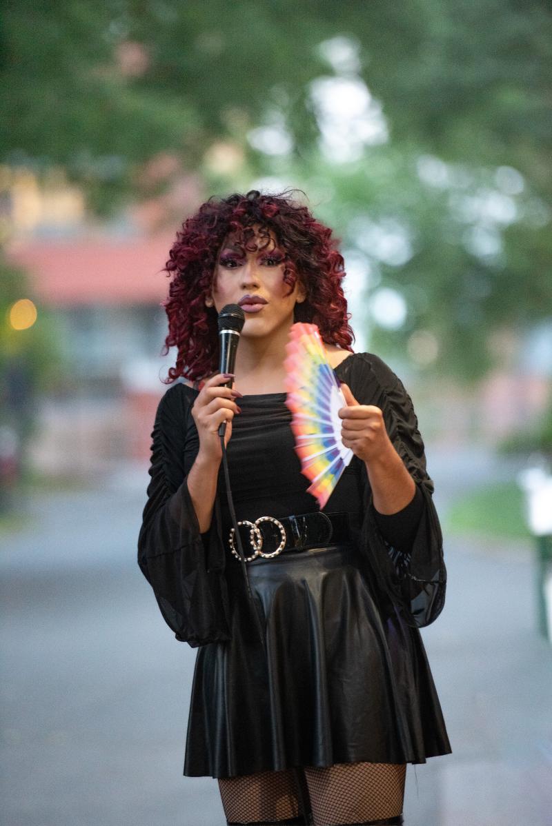 A person with curly long dark red hair holds a mic and a rainbow fan