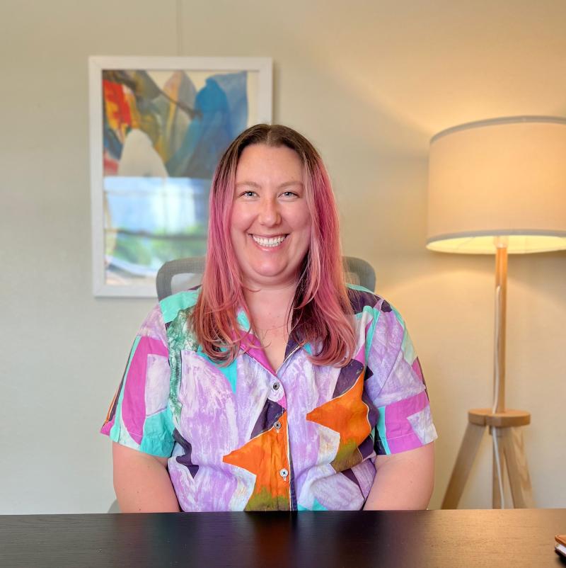 Ellie Lipton smiles at the camera wearing a colorful button down shirt and sitting at a table. There is a lamp behind them and a painting.