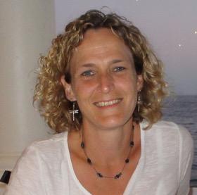 Yvette Esprey who has medium length light hair wearing a white shirt smiles in front of an ocean