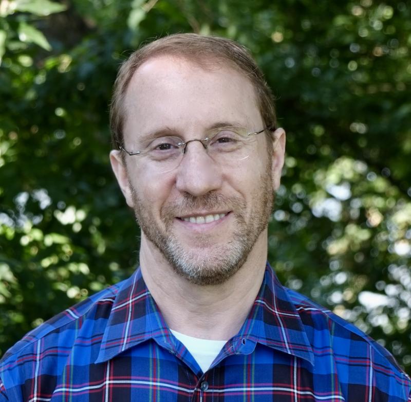 Instructor Kounitz stands in front of greenery in a blue, black, and white plaid button-up, smiling with glasses.