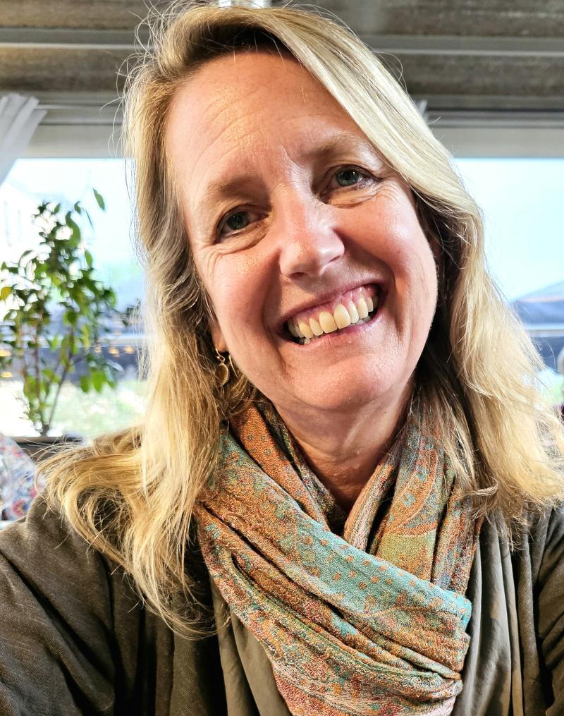 instructor lisa howard smiles with head tilted in front of a window wearing a green and brown scarf