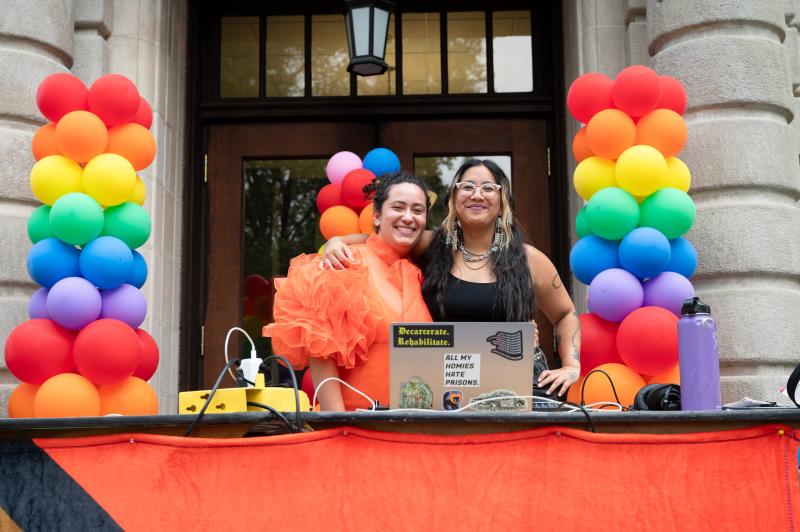 Student deejays at 2023 Pride celebration stand behind a table with a laptop on it and rainbow balloon sculptures on either side.