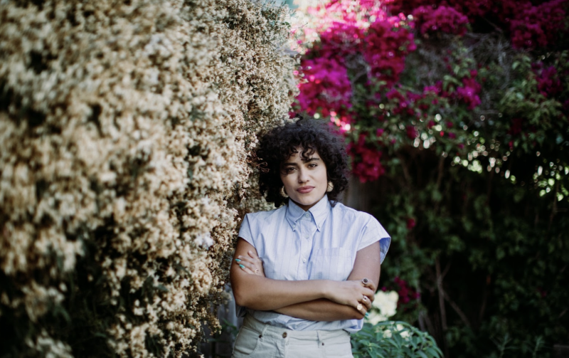 Jess Semaan, a person with curly dark hair wearing a white shirt stands with arms crossed against a flowering bush with white blossoms with pink blossoms behind