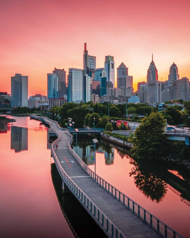 City of Philadelphia- grey skyscrapers, a bridge in the foreground, with pink sky and pink reflection on the water in the foreground