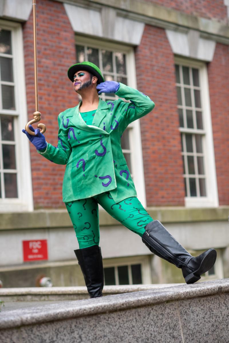 Leo Crayz, wearing green suit covered with purple question marks, standing outside of a brick building wearing a bowler and holding a golden cane