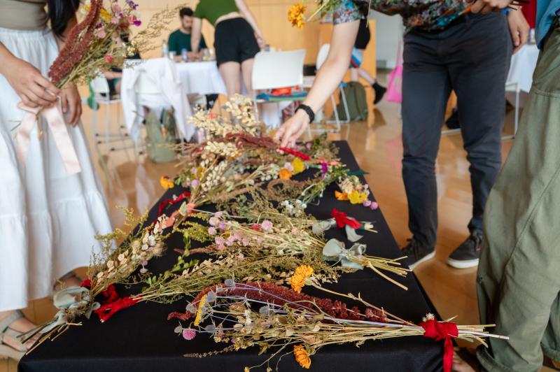 From Closing Ceremony 2025, a table with black table cloth covered in dried flower bouquets with people's lower bodies visible as they lay flower down