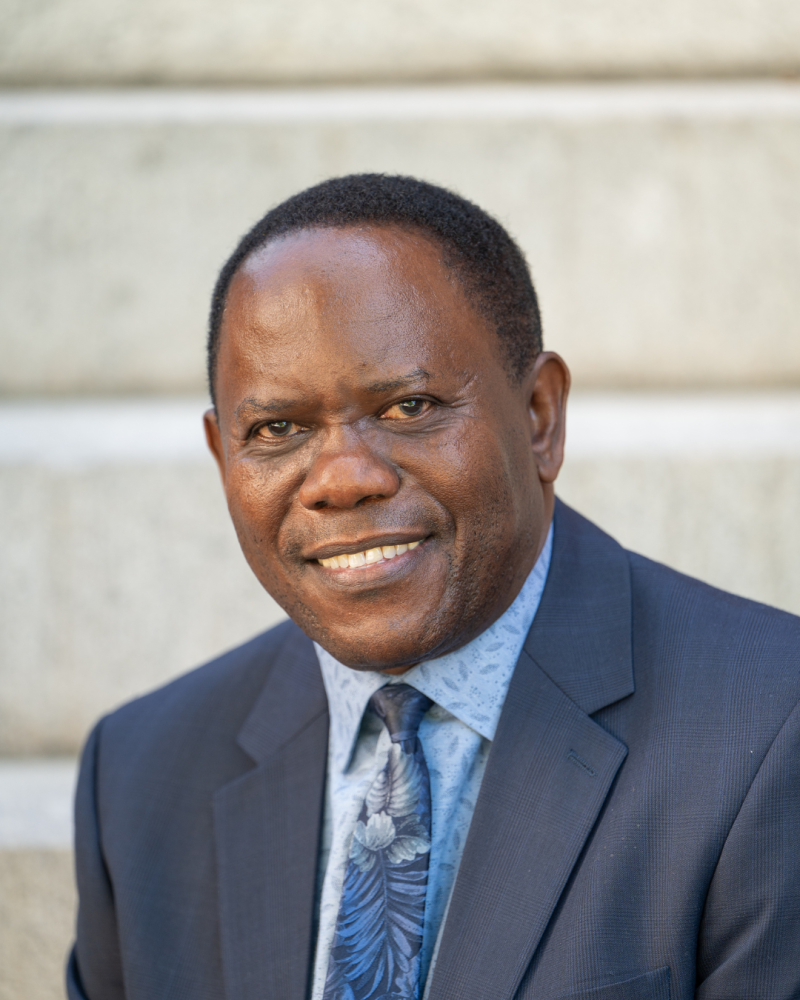 Hugo Kamya smiling in blue suit and tie with grey cement wall behind