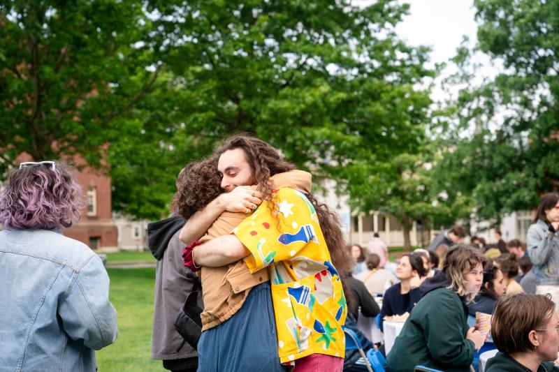 Two students hugging at opening ceremony 2025. People sitting at tables behind, green trees and brick building in background