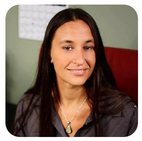 Jus Crea headshot, a person with long dark hair sitting with green wall behind