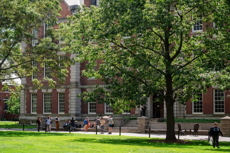 Students sitting on a cement bench in front of a brick building, green grass, green trees