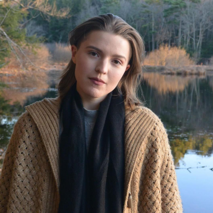 Leah Eve-Cowles stands in front of a lake wearing a brown sweater and black scarf.