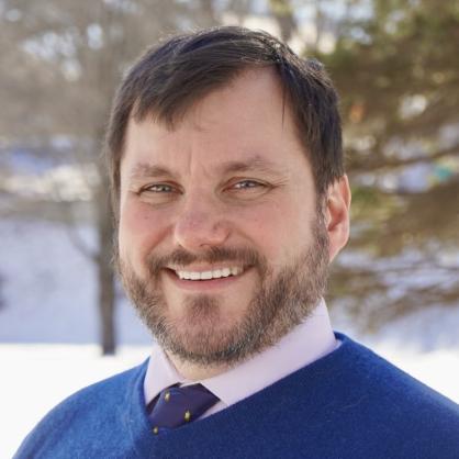 Kurt White wearing blue sweater and tie with snow and trees behind