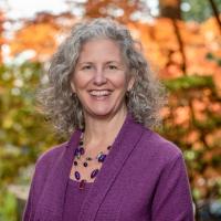 Mary Curtin smiles at the camera. She is wearing a purple sweater and has shoulder length curly hair.