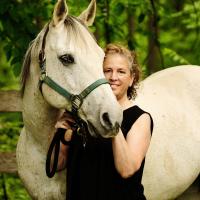 Dana Spett standing cheek to cheek with a flea-bitten grey horse wearing a green halter. Behind is a wooden fence and greenery