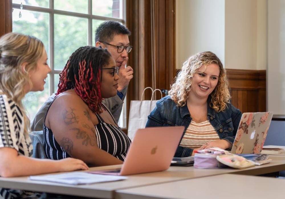 Three students sit in a classroom with laptops while a faculty member stands nearby engaged in conversation.
