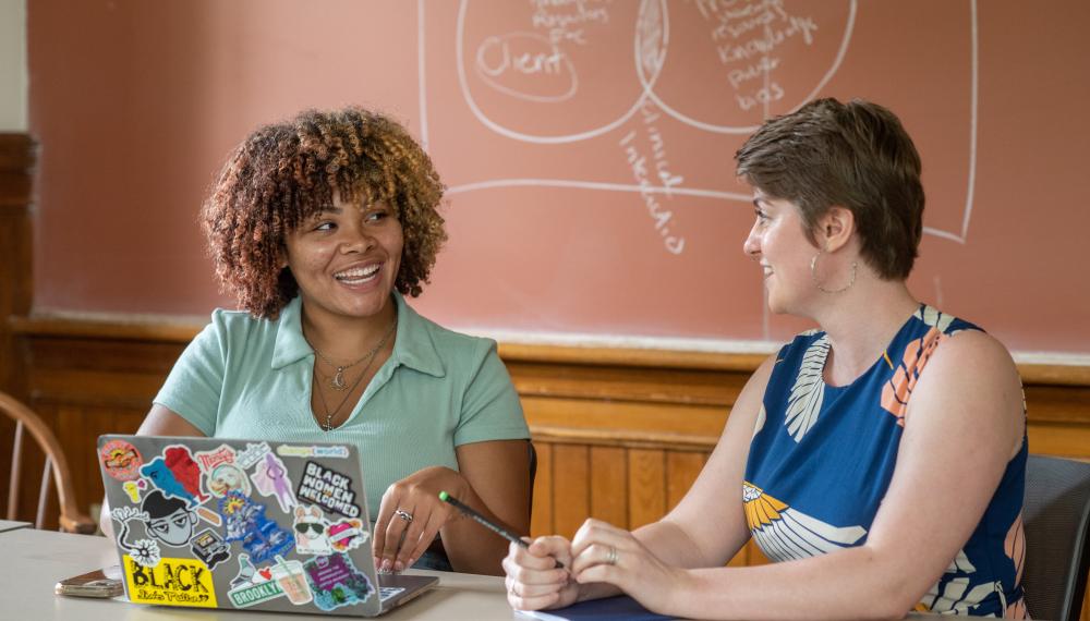 Two students sit at a table engaged in conversation. There is a laptop on the table in front of them.