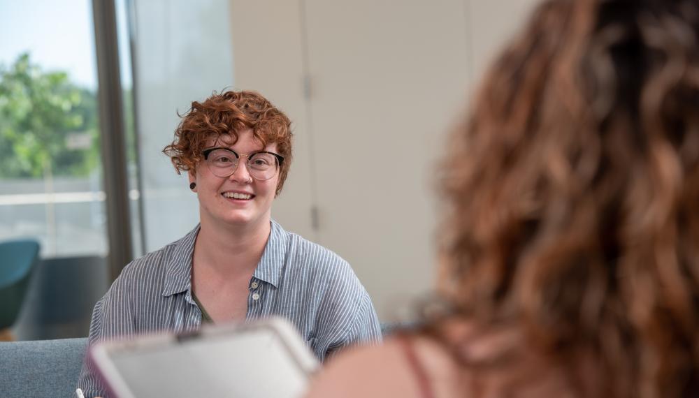Photo of Taylor Haaf wearing a blue and white striped button up. They have short curly reddish hair and glasses