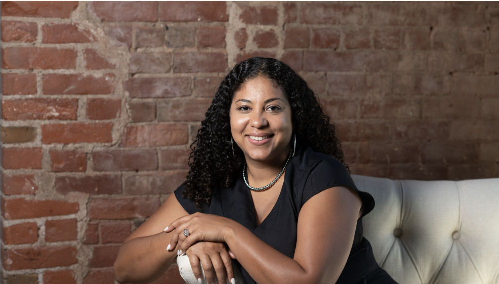Photo of a person with long curly dark hair sitting on a white tufted couch. They are wearing a black v-neck t-shirt and have their arms resting on the arm of the sofa.