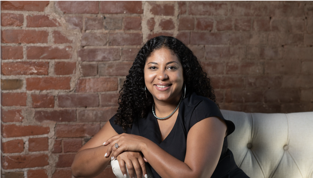 Photo of a person with long curly dark hair sitting on a white tufted couch. They are wearing a black v-neck t-shirt and have their arms resting on the arm of the sofa. 